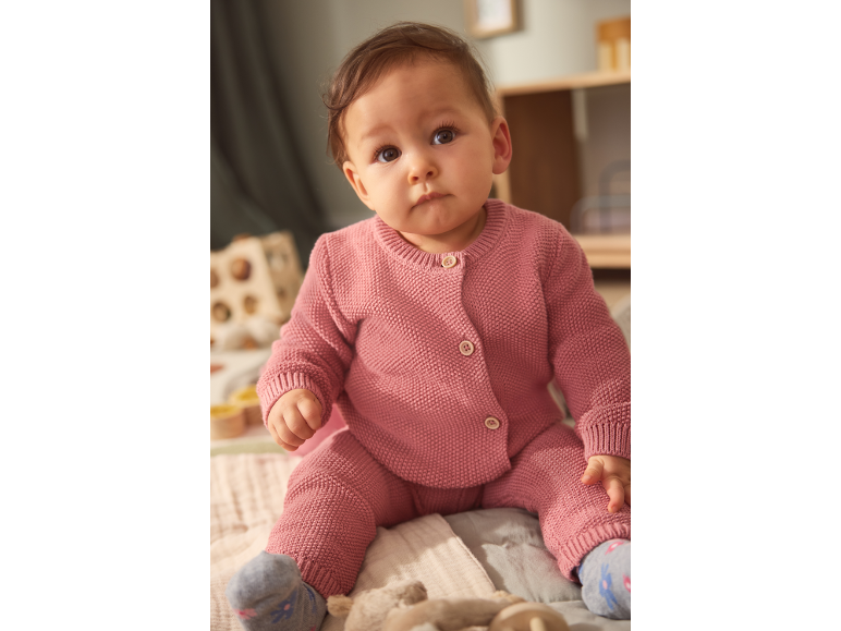 Baby in a pink knitted set, sitting on a blanket.