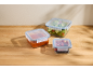 Three glass food containers with blue lids, filled with salad, sauce, and berries, on a wooden table.
