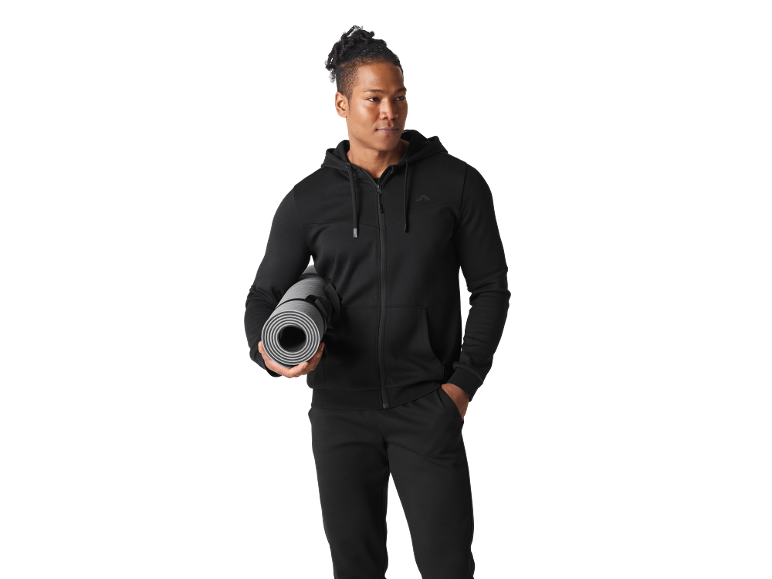 Man in black tracksuit holding a rolled-up yoga mat.
