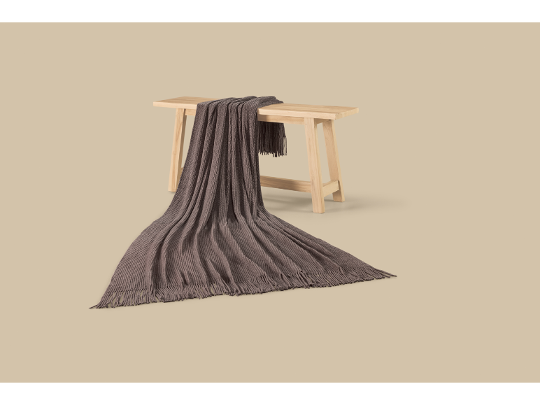 Dark brown knitted blanket with fringes draped over a light wooden bench.