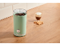 Green coffee grinder with coffee beans, latte coffee, and cookies on a wooden table.