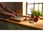 Hands cutting herbs on a wooden chopping board, next to spices and potted herbs.