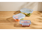 Three glass food containers with blue plastic lids, filled with vegetables and pasta, on a wooden table.