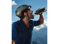 Man in a cycling helmet and sports shirt drinking from a bottle, with mountains in the background.