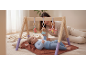 Baby playing with a wooden activity gym with hanging toys, smiling woman in the background.