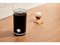 Coffee grinder with beans, a glass of coffee, and cookies on a wooden counter.