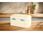 Cream bread bin on a wooden kitchen counter with spices and a plant in the background.