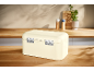 Cream bread bin on a wooden kitchen counter with spices and a plant in the background.