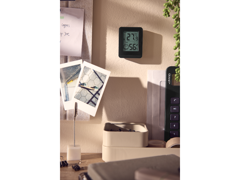 Digital thermometer displaying 27.9°C and 56% humidity on a wall, next to notes and photos.