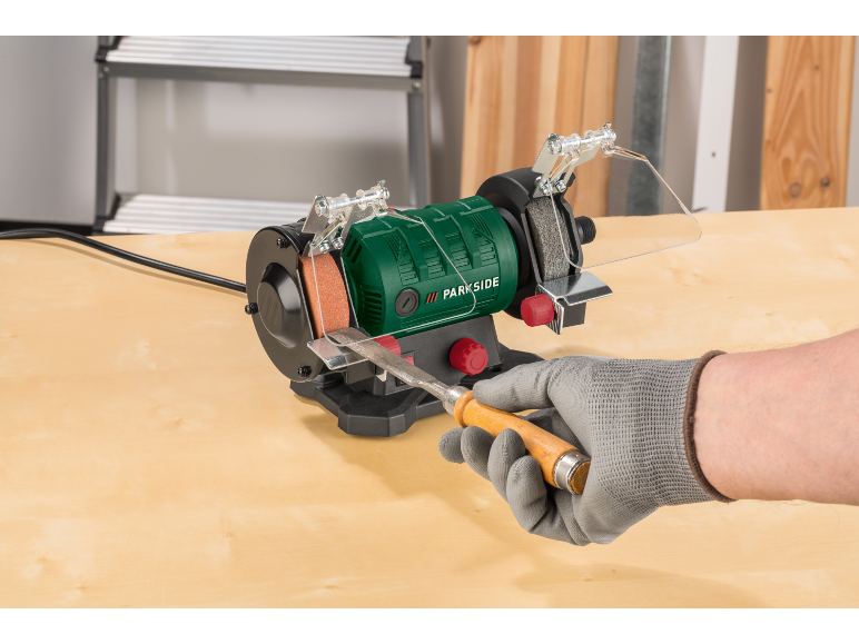 Green bench grinder with a wood chisel being sharpened by a gloved hand.