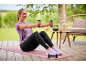 Woman exercising outdoors on a pink mat, holding dumbbells, wearing a lilac t-shirt and black leggings.