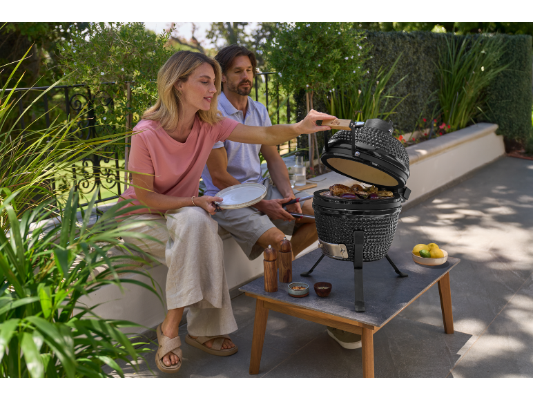Couple grilling meat and vegetables on a ceramic table barbecue on a patio.