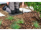 Gardener kneeling on a kneeling pad holding a trowel, surrounded by mulch and plants.