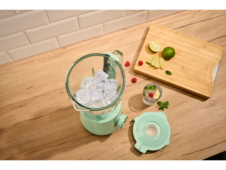 Mint-colored blender with ice cubes, limes, raspberries, and mint on a wooden table.