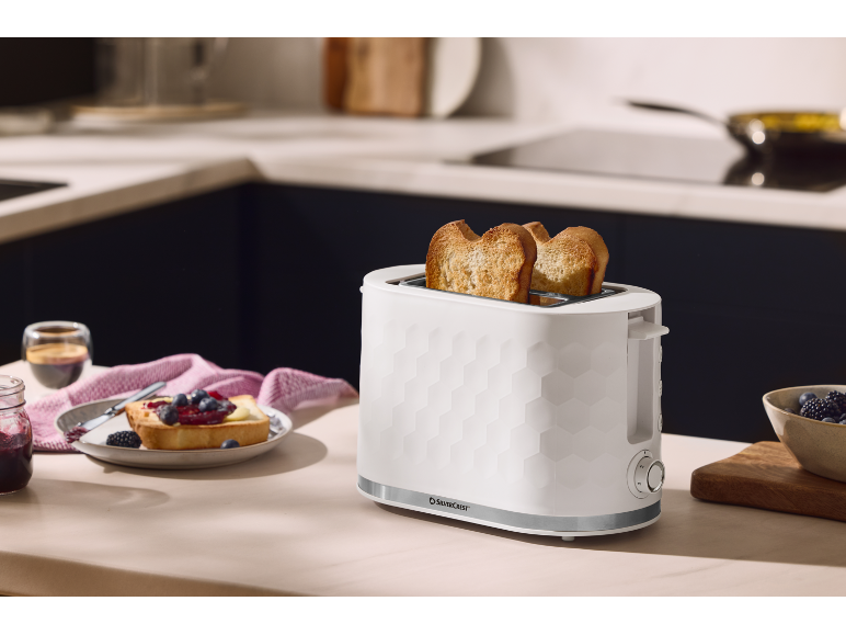 White toaster with toast on a kitchen counter, next to a plate with jam and fruit.
