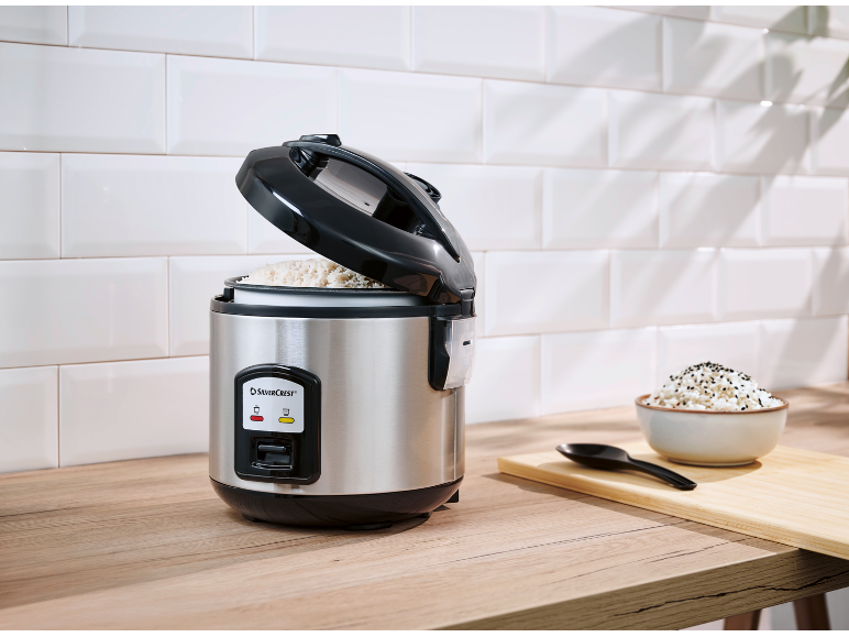 Open rice cooker with rice, a bowl of rice, and a spoon on a wooden countertop.