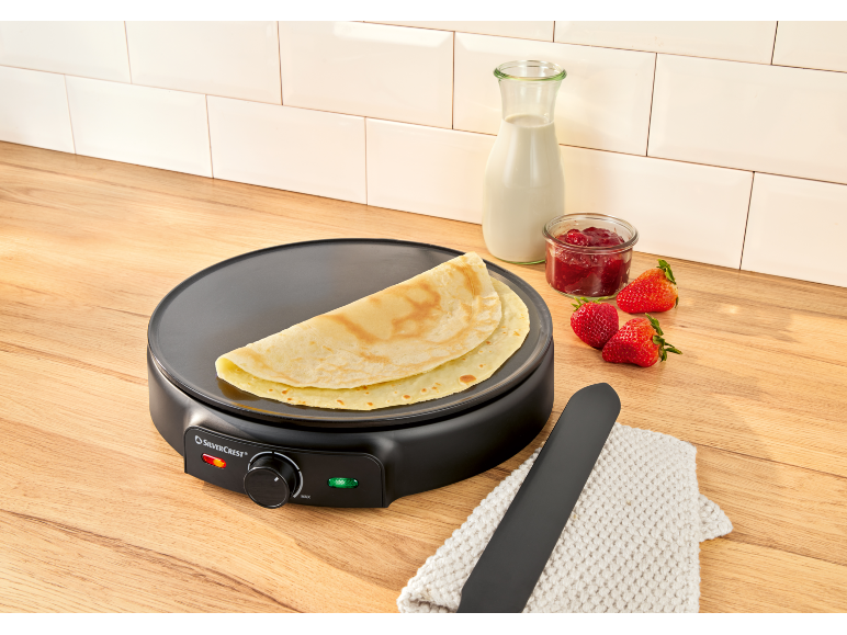 Electric crepe maker with crepes, milk, jam, and strawberries on a wooden countertop.