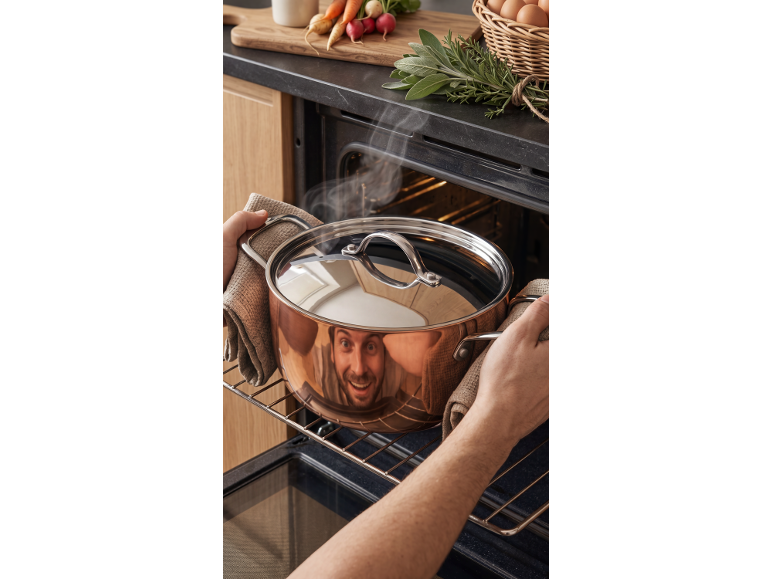 Man putting a pot with steam into an oven, with fresh vegetables and herbs nearby.