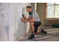 Man in shorts and grey t-shirt, kneeling, removing wallpaper with a scraper.