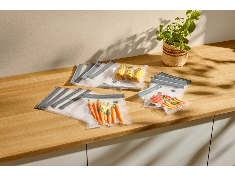 Vacuum bags with carrots, corn, and apples on a wooden countertop.