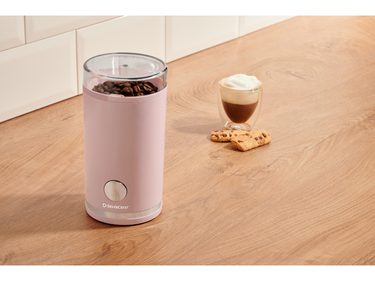 Pink coffee grinder with coffee beans, coffee drink, and cookies on a wooden surface.