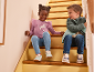 Two children in casual wear and sneakers sitting on wooden stairs, smiling.