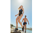 Woman in black swimsuit and man in swim shorts walking on rocks by the sea.