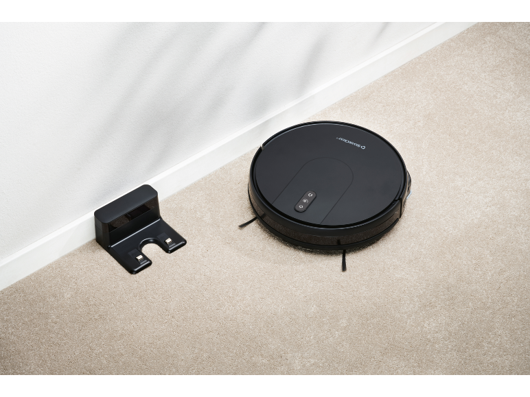 Black robotic vacuum cleaner next to its charging station on a beige carpet.