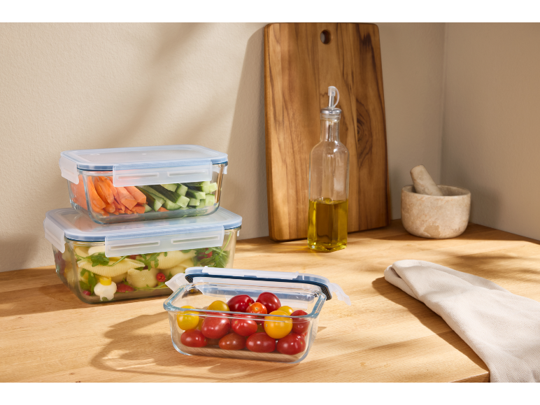 Glass food storage containers with vegetables and pasta on a wooden countertop.