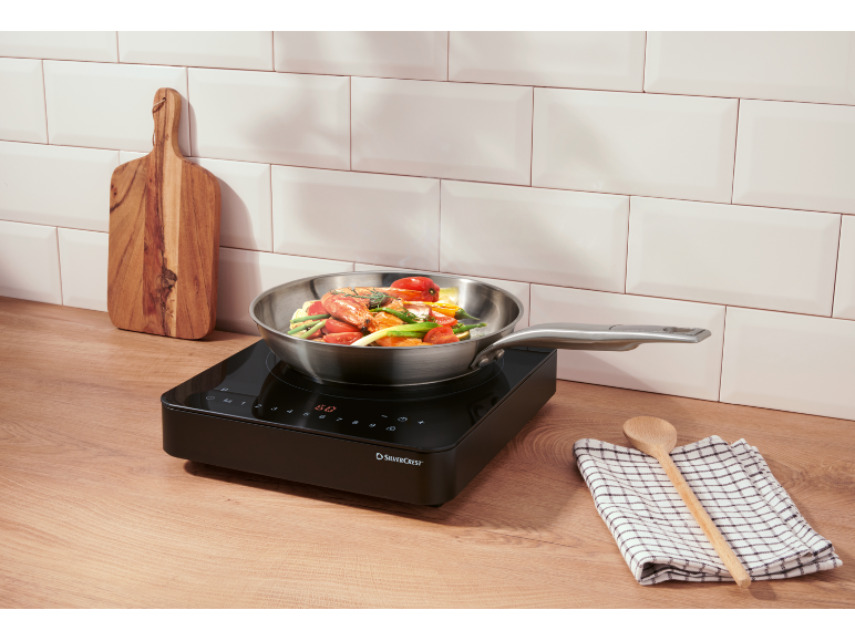 Induction hob with vegetables and seafood cooking on a wooden countertop.