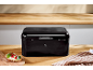 Glossy black bread bin on a wooden countertop with a sandwich and a kitchen towel.