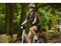 Woman on a bicycle with a backpack, helmet, and sunglasses in a forest setting.