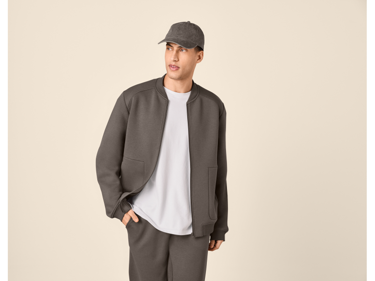 Man in a dark grey bomber jacket, white t-shirt, and matching sweatpants with a cap