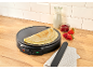Electric crepe maker with crepes, milk, jam, and strawberries on a wooden countertop.