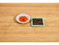 Diced red bell pepper on a white plate next to a vegetable chopper on a wooden surface.