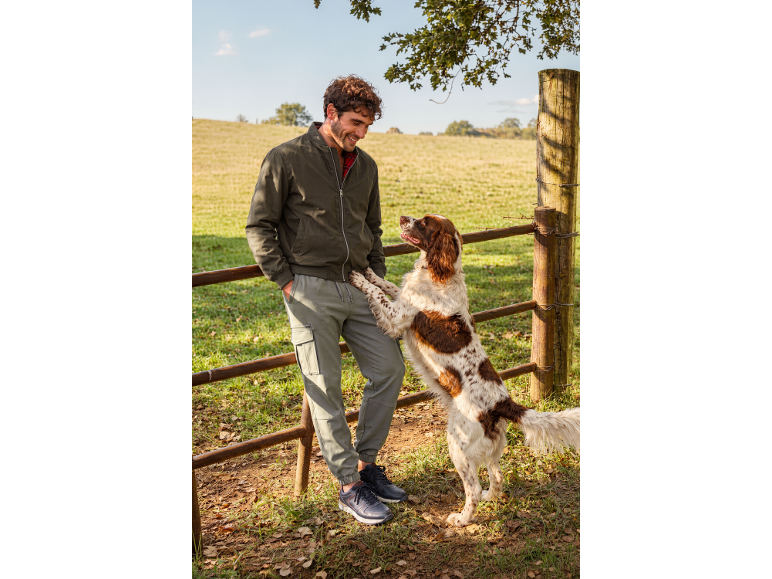 Man in bomber jacket and cargo pants with a dog in a field.