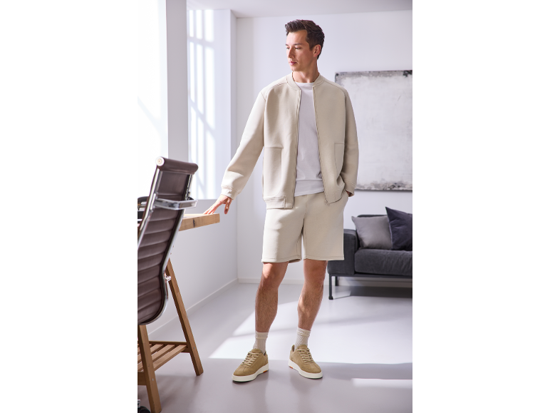 Man in a beige bomber jacket, shorts, and sneakers, posing in a bright office.