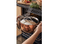 Man putting a pot with steam into an oven, with fresh vegetables and herbs nearby.