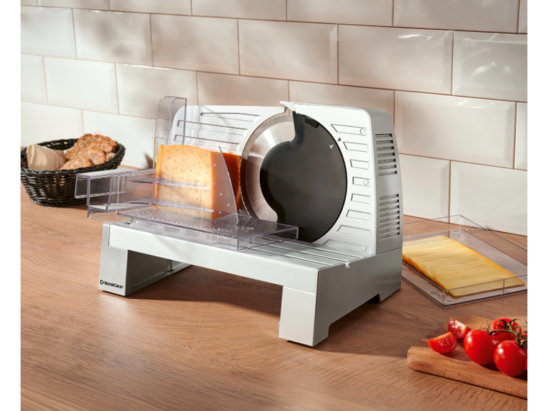 Electric food slicer with cheese, bread, and tomatoes on a wooden kitchen counter.