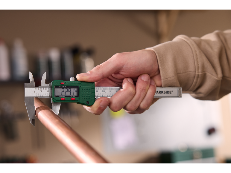 A person measures a copper pipe with a digital caliper, showing 22.91 mm on the display.