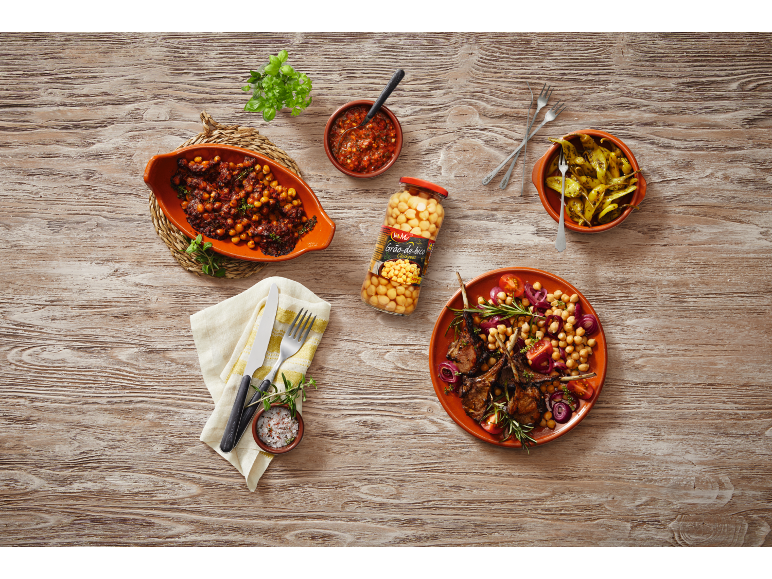 Chickpea dish with lamb chops, peppers, and sauce on a wooden table.