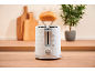 White toaster with a bread roll on top in a kitchen with cutting boards.