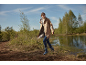 Woman in a beige coat and blue boots walking in nature by the water.