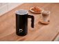 Black milk frother on a wooden table with a latte and croissants in the background.