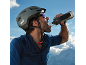 Man in helmet and sportswear drinking water from a bottle, with mountains in the background.