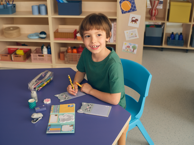 Smiling boy writing in a cat-shaped notebook, with cat-themed school supplies.