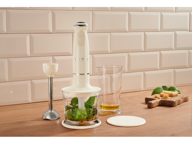 Cream hand blender with accessories, herbs, oil, and cheese on a wooden countertop.