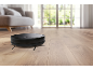 Black robotic vacuum cleaner on a wooden floor in a bright living room.