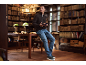 Man in a sweater and jeans sitting on a table in a library, reading a book.