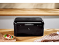 Glossy black bread bin on a wooden countertop with breakfast on the side.
